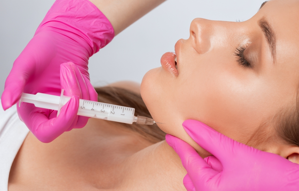 Woman receiving facial injection in jaw area by gloved hands, eyes closed.