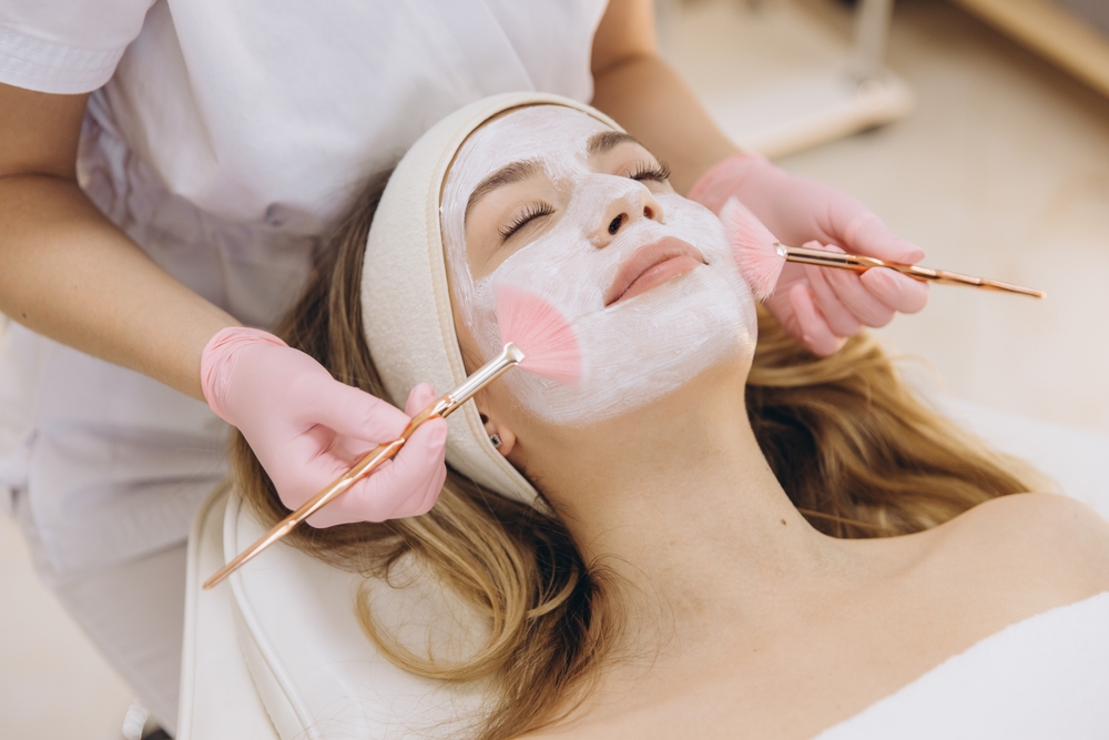 Woman receiving facial treatment with brushes, wearing a headband and white mask, attended by a professional in gloves.