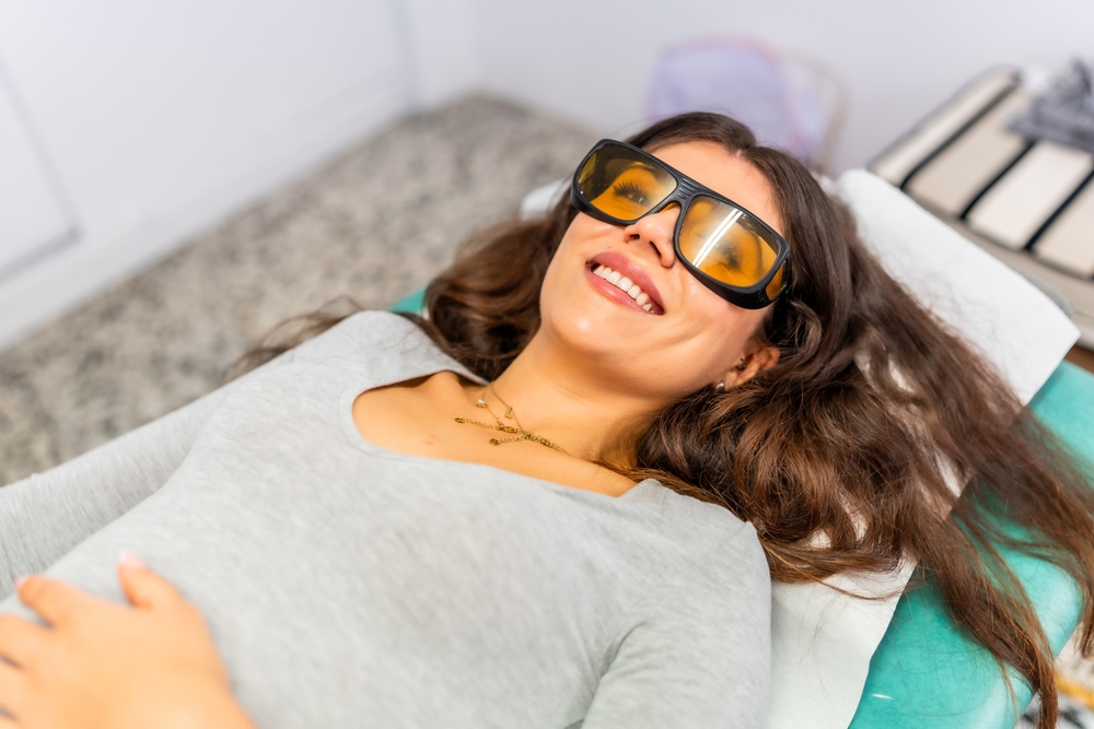 Woman smiling, lying on a treatment table, wearing protective goggles, in a clinical setting.
