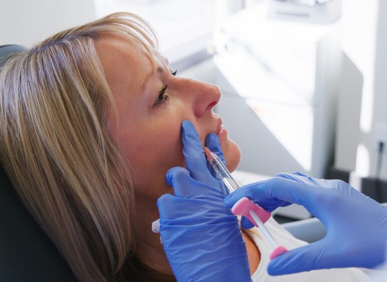 Woman receiving cheek filler injection from gloved professional in a clinical setting.