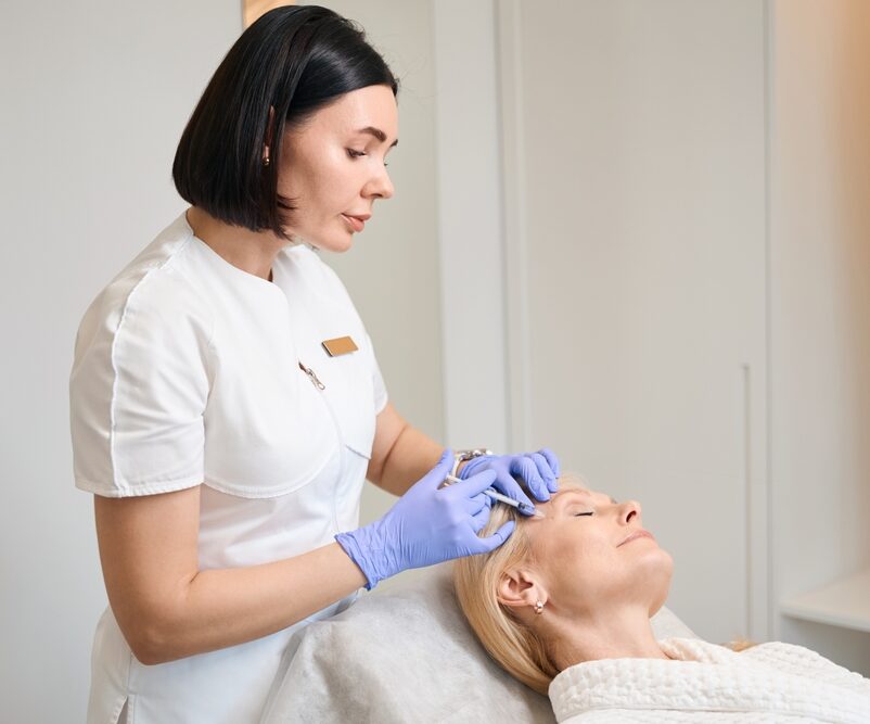 Aesthetician in white uniform and gloves administering temple filler to a reclining woman.