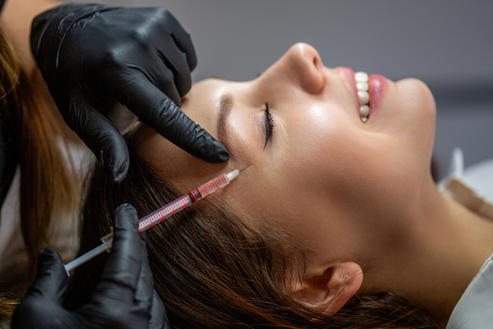 Woman receiving temple filler injection from gloved professional, smiling and relaxed.