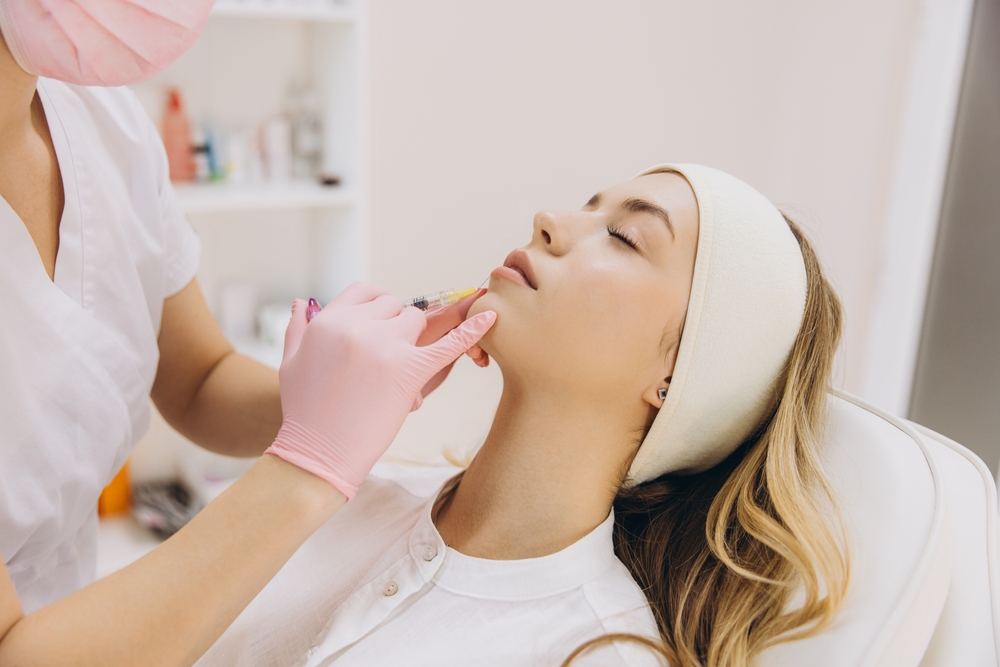 Aesthetician in pink gloves administering lip filler injection to a woman lying back with a headband.