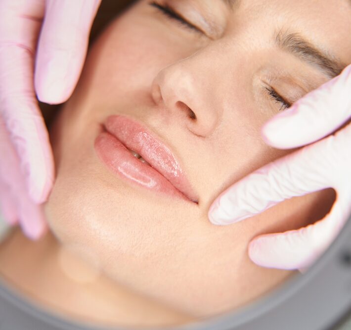 Close-up of a person's face with glossy lips, eyes closed, and gloved hands gently touching the cheeks.
