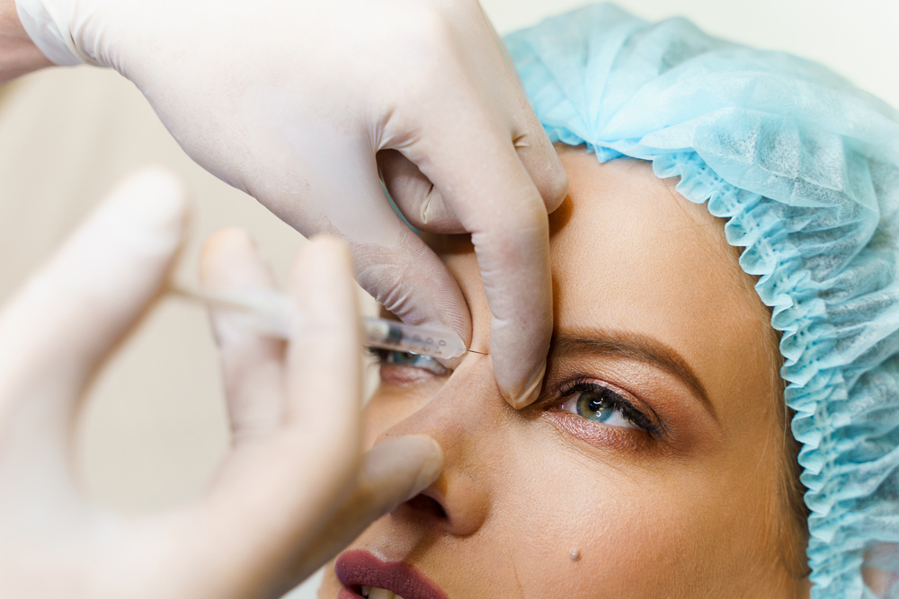Person receiving a cosmetic injection in the nose, wearing a blue surgical cap, close-up view.